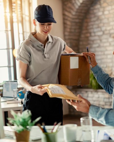 Smiling female courier delivering package to a businessman while he is working in the office.